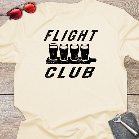 A cream-colored t-shirt with a black distressed graphic reading 'FLIGHT CLUB' above and below four pint glasses on a tray, laid flat on a wood surface with sunglasses, a bottle opener, and a coaster as props.
