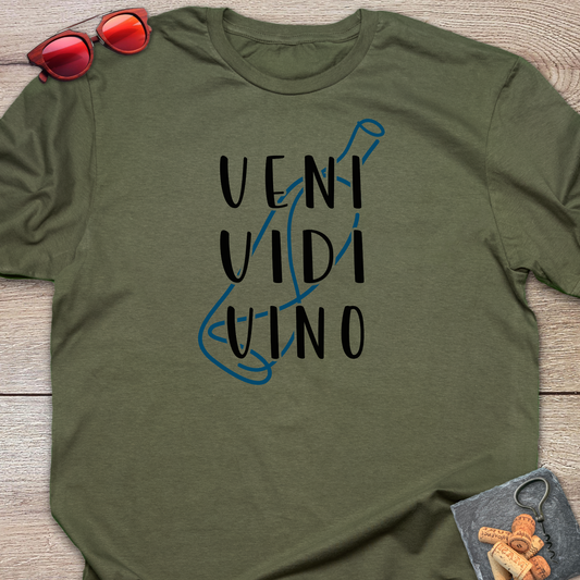 An olive green crewneck t-shirt with a graphic print reading 'VENI VIDI VINO' in black over a blue wine bottle outline, styled flat on a wooden surface with red sunglasses and a small corkscrew and corks in the corners.