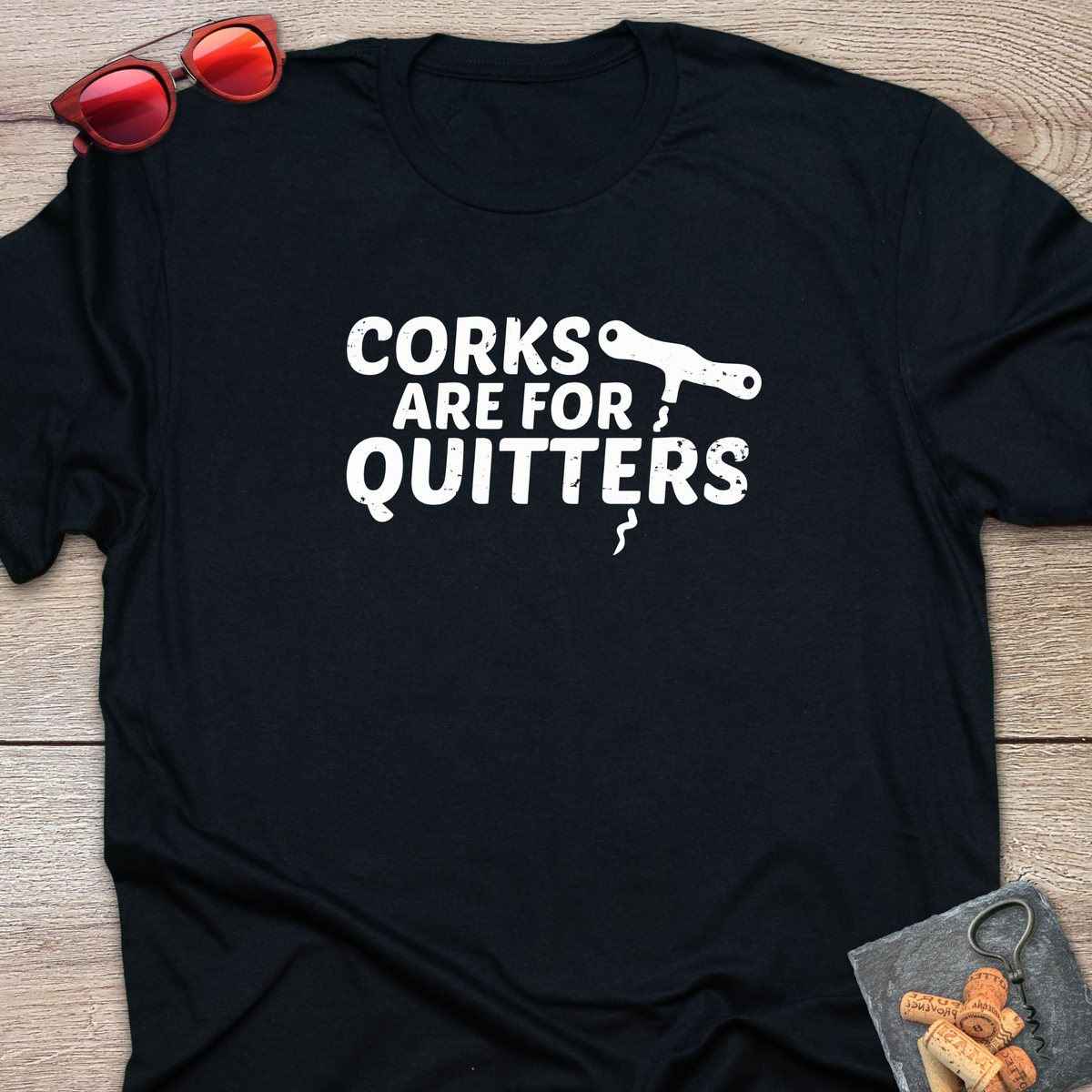 An black t-shirt with a distressed white coloured graphic that reads 'CORKS ARE FOR QUITTERS' alongside a small cork driver tool illustration. The t-shirt is laid flat on a light wood background, styled with red sunglasses in the top-left corner and a small corkscrew with corks in the bottom-right corner.