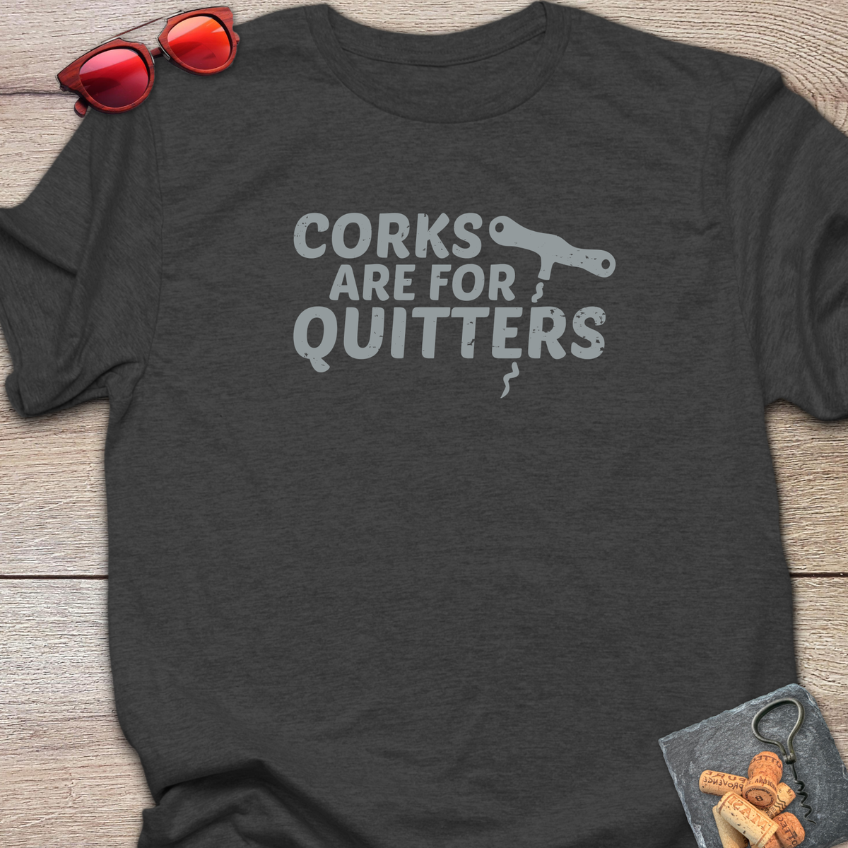 An dark heather  t-shirt with a distressed light gray coloured graphic that reads 'CORKS ARE FOR QUITTERS' alongside a small cork driver tool illustration. The t-shirt is laid flat on a light wood background, styled with red sunglasses in the top-left corner and a small corkscrew with corks in the bottom-right corner.