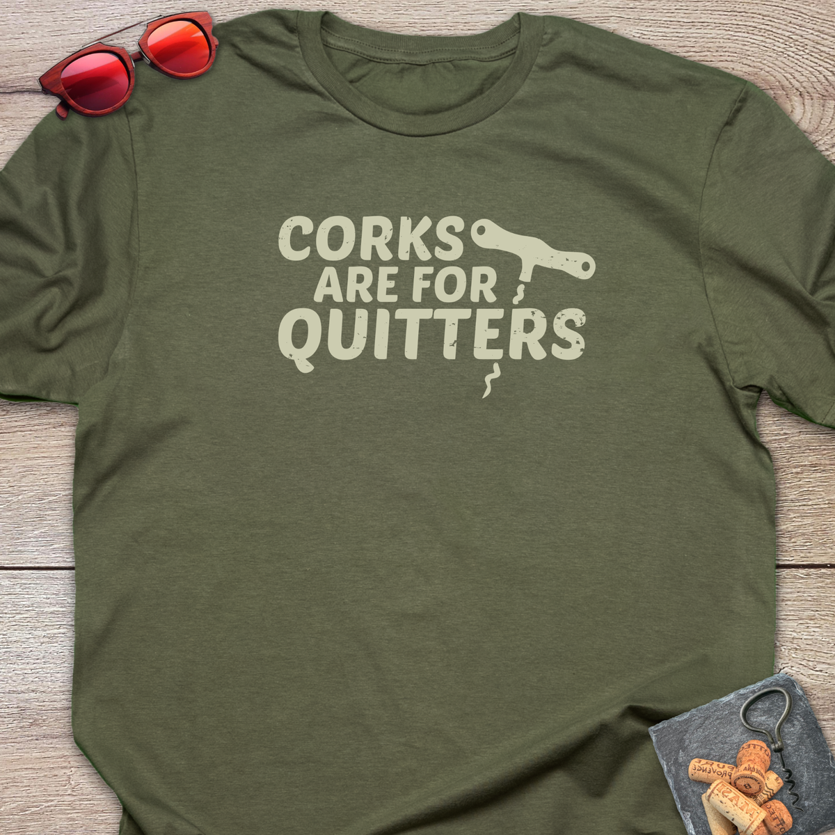 An olive green t-shirt with a distressed light green coloured graphic that reads 'CORKS ARE FOR QUITTERS' alongside a small cork driver tool illustration. The t-shirt is laid flat on a light wood background, styled with red sunglasses in the top-left corner and a small corkscrew with corks in the bottom-right corner.
