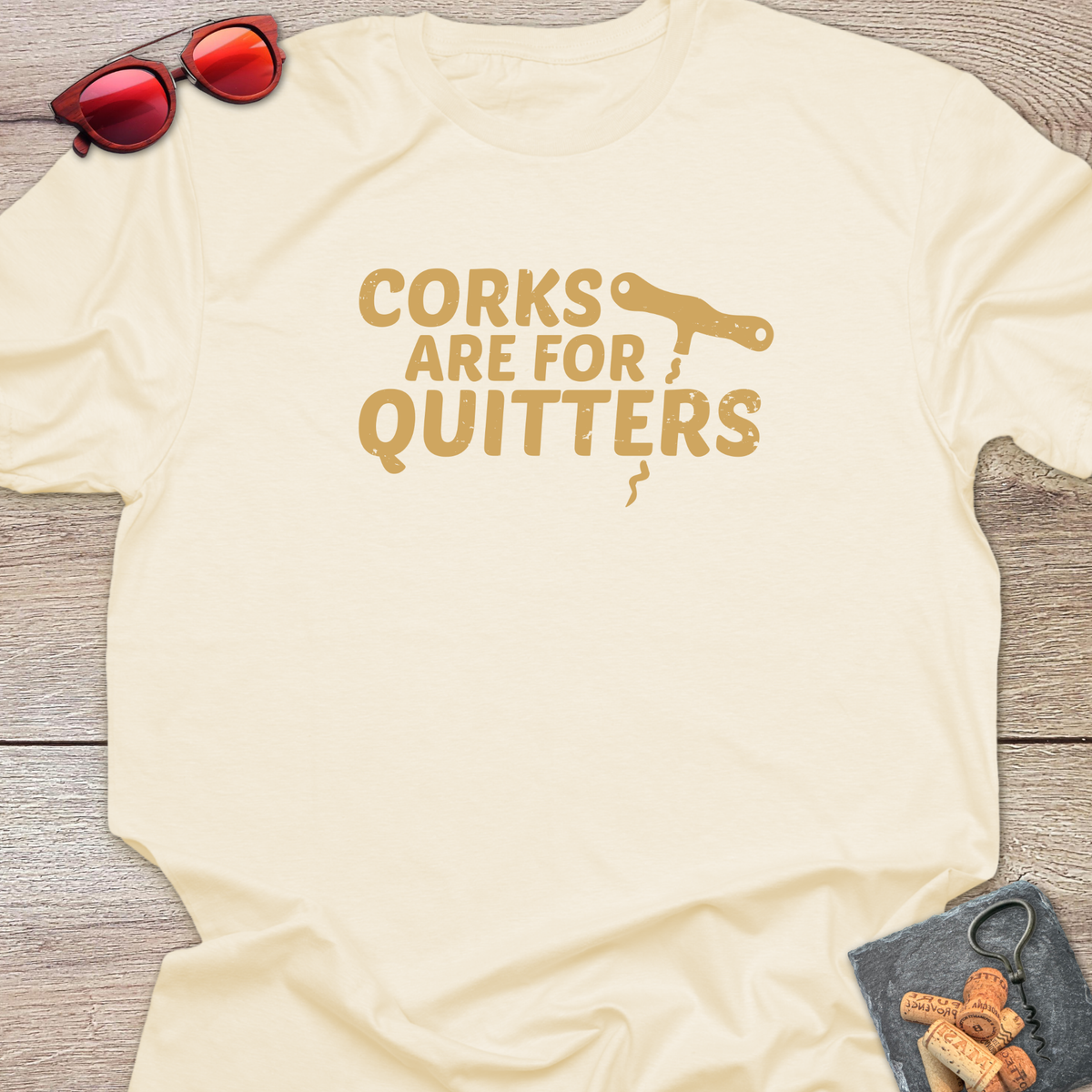 An natural  t-shirt with a distressed tan coloured graphic that reads 'CORKS ARE FOR QUITTERS' alongside a small cork driver tool illustration. The t-shirt is laid flat on a light wood background, styled with red sunglasses in the top-left corner and a small corkscrew with corks in the bottom-right corner.