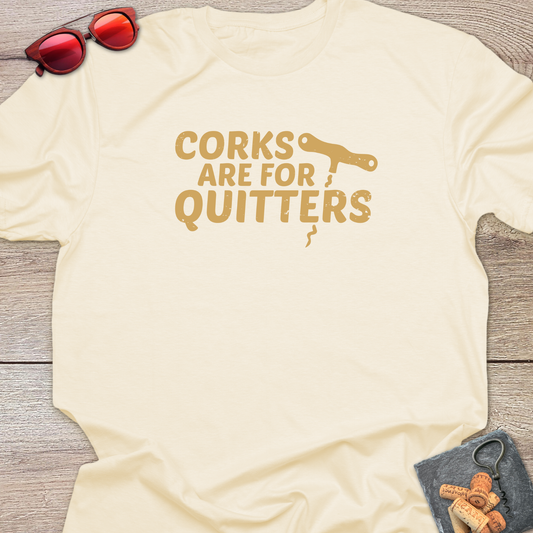 An natural  t-shirt with a distressed tan coloured graphic that reads 'CORKS ARE FOR QUITTERS' alongside a small cork driver tool illustration. The t-shirt is laid flat on a light wood background, styled with red sunglasses in the top-left corner and a small corkscrew with corks in the bottom-right corner.