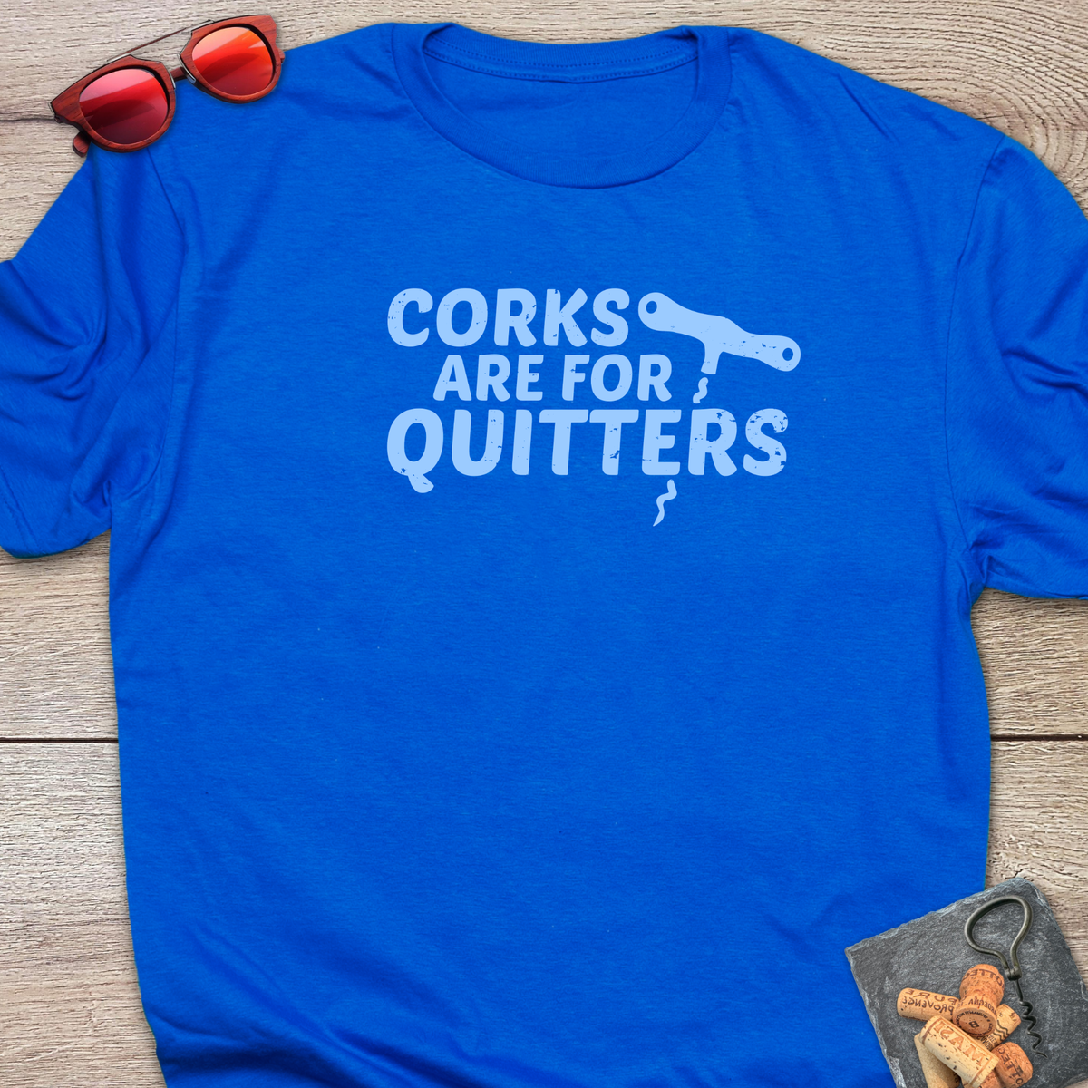 An royal blue t-shirt with a distressed light blue coloured graphic that reads 'CORKS ARE FOR QUITTERS' alongside a small cork driver tool illustration. The t-shirt is laid flat on a light wood background, styled with red sunglasses in the top-left corner and a small corkscrew with corks in the bottom-right corner.