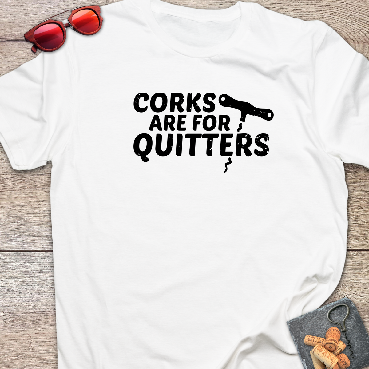 An white t-shirt with a distressed black coloured graphic that reads 'CORKS ARE FOR QUITTERS' alongside a small cork driver tool illustration. The t-shirt is laid flat on a light wood background, styled with red sunglasses in the top-left corner and a small corkscrew with corks in the bottom-right corner.