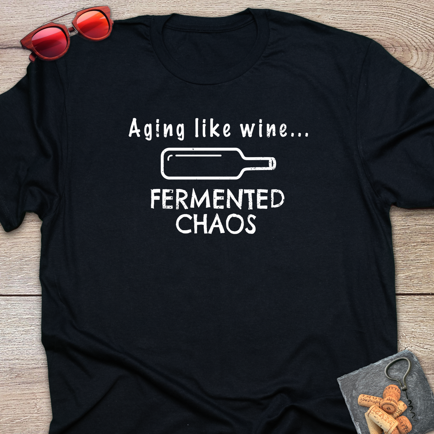 A black short-sleeve graphic T-shirt laid flat on a wood surface, with white text reading 'Aging like wine. Fermented Chaos.' And a graphic of wine bottle in between the two statements.