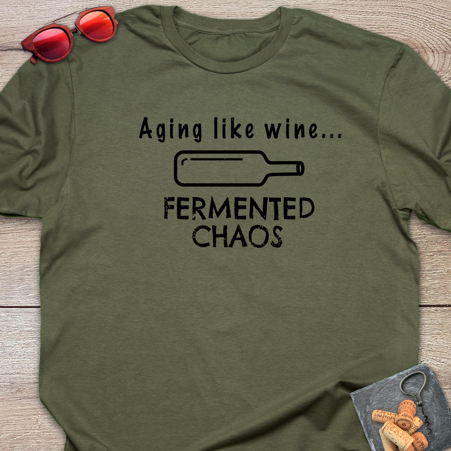 An olive green crewneck t-shirt with a black graphic print that reads 'Aging like wine. Fermented Chaos.' And a graphic of wine bottle in between the two statements.