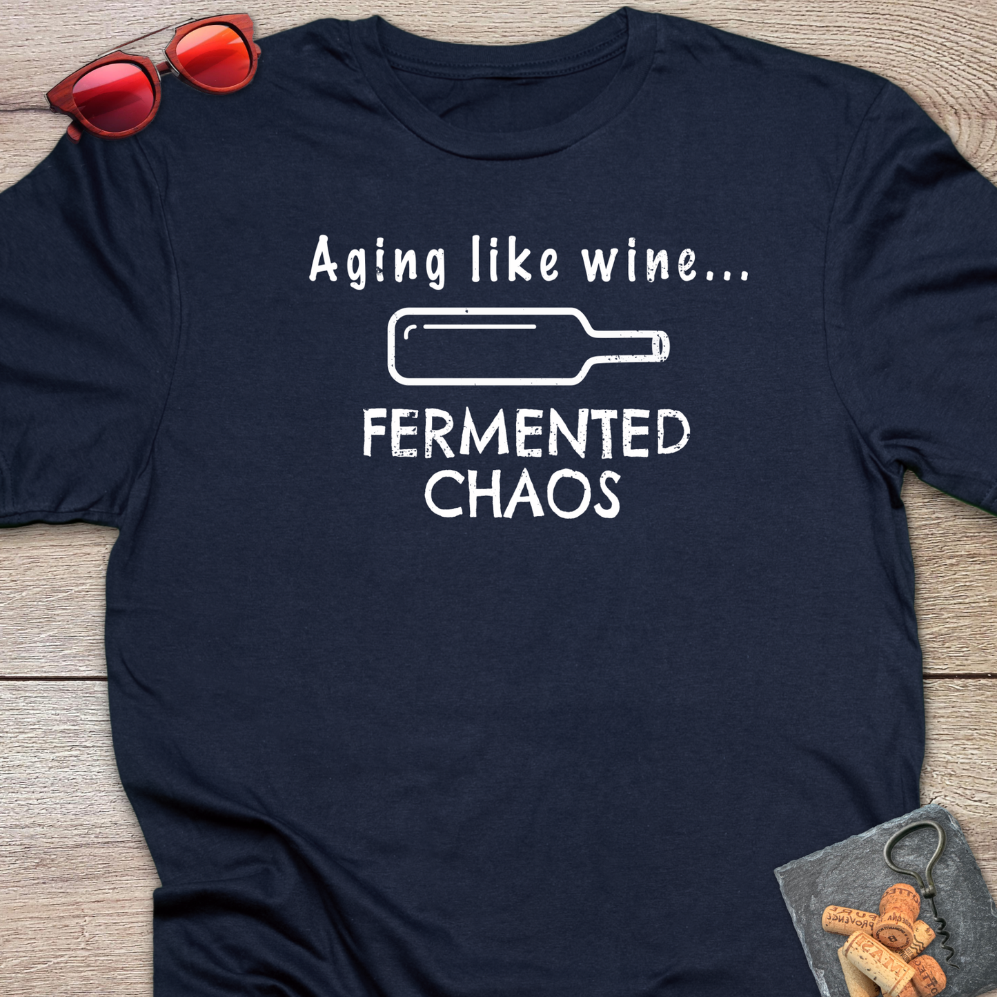 A navy blue graphic t-shirt laid flat on a wooden surface, featuring the white text 'Aging like wine. Fermented Chaos.' And a graphic of wine bottle in between the two statements.