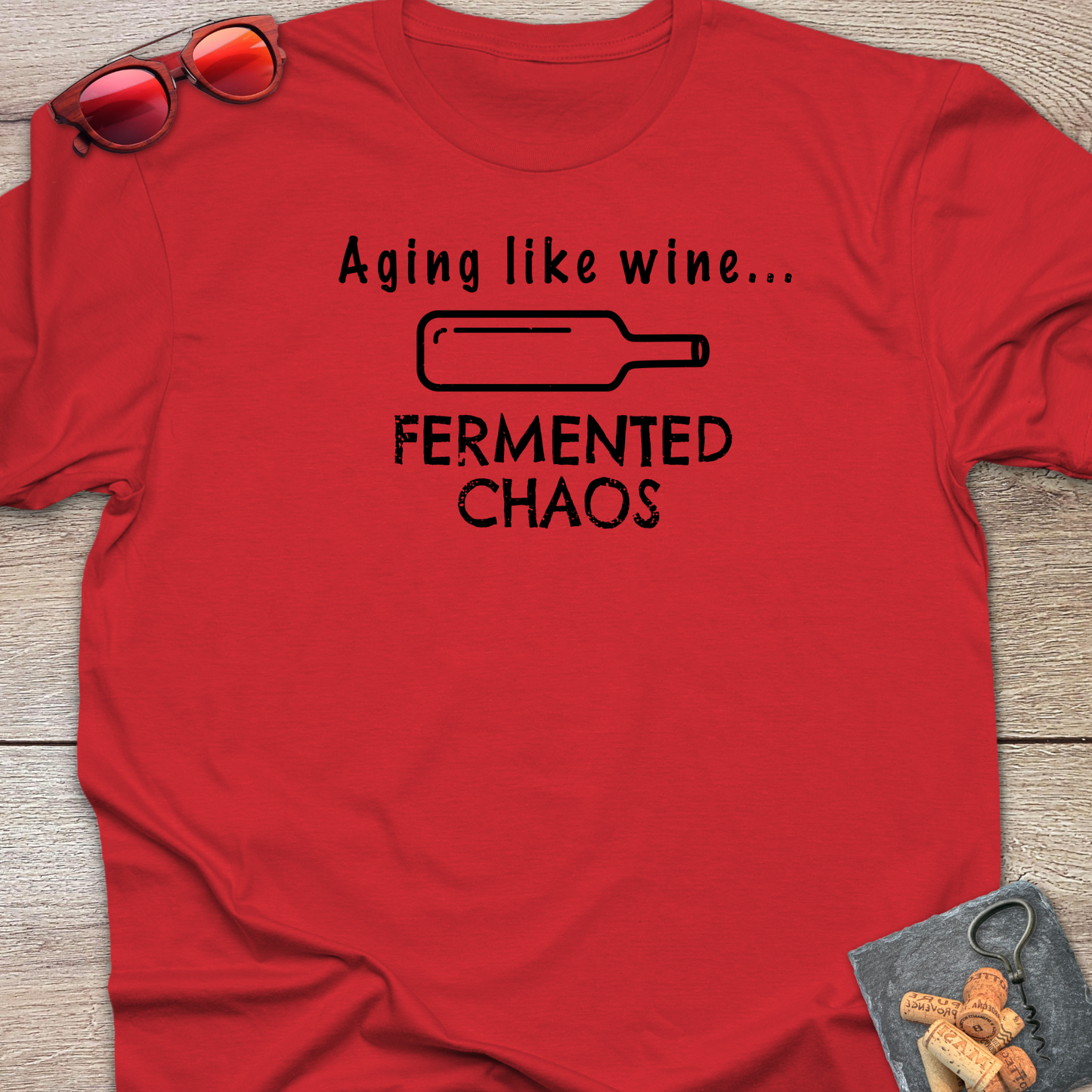 A red short-sleeve graphic T-shirt laid flat on a wood surface, with black text reading 'Aging like wine. Fermented Chaos.' And a graphic of wine bottle in between the two statements.