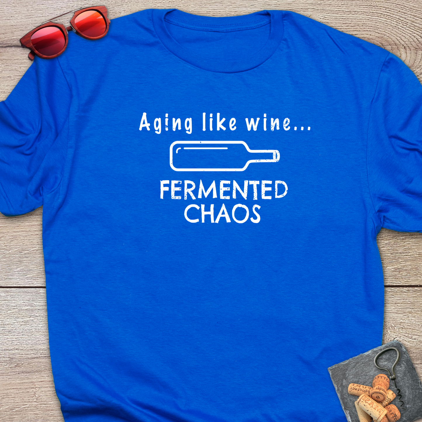 A royal blue short-sleeve t-shirt laid flat on a rustic wood background, featuring a white graphic that reads 'Aging like wine. Fermented Chaos.' And a graphic of wine bottle in between the two statements.