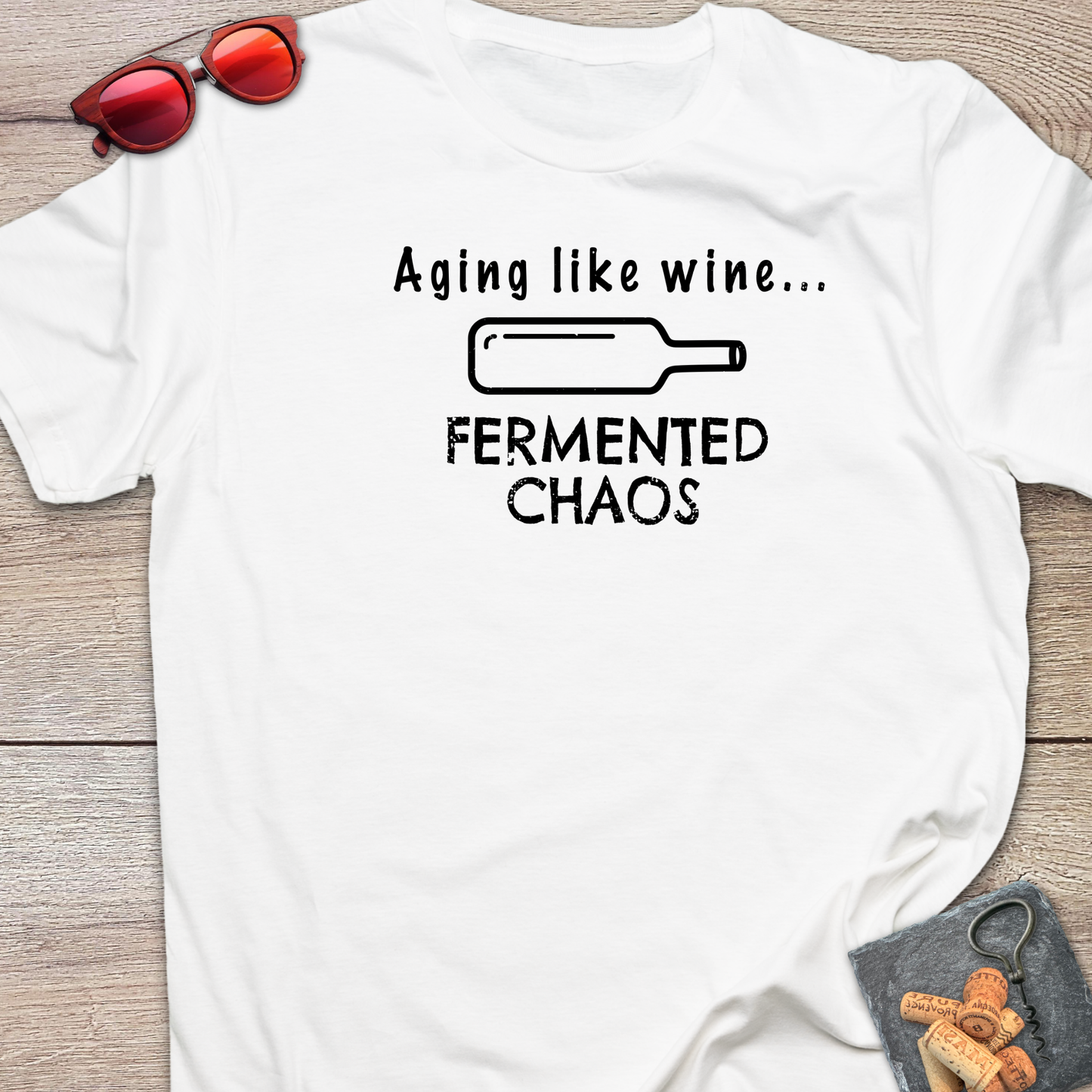 A white short-sleeve graphic T-shirt laid flat on a wood surface, with black text reading 'Aging like wine. Fermented Chaos.' And a graphic of wine bottle in between the two statements.