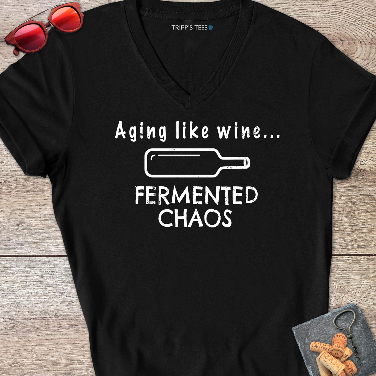 A black v-neck t-shirt with a white distressed graphic reading 'Aging like wine...Fermented Chaos' and a graphic of a wine bottle on it's side, laid flat on a wood surface with sunglasses, wine opener and corks on a coaster. 