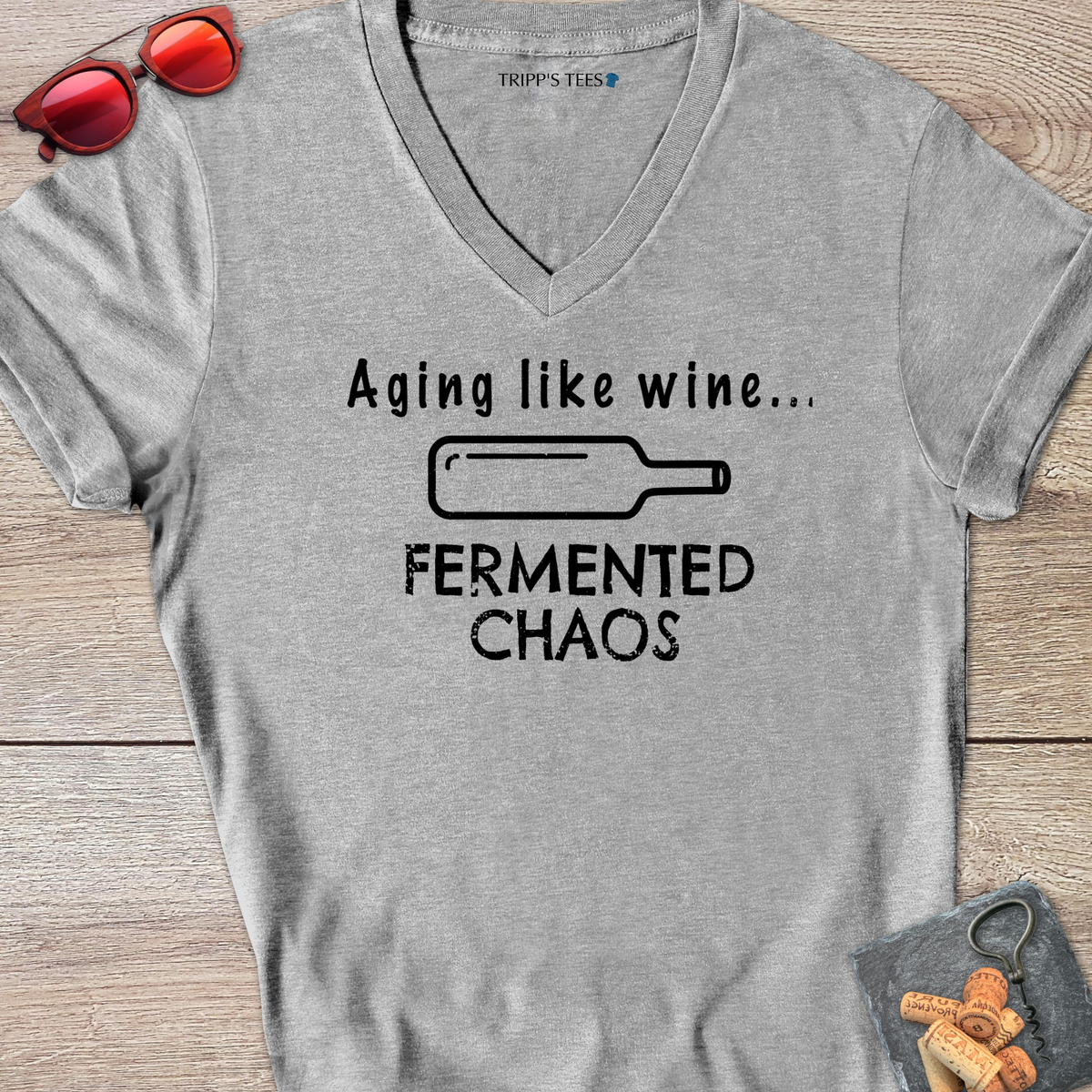 A sport gray v-neck t-shirt with a black distressed graphic reading 'Aging like wine...Fermented Chaos' and a graphic of a wine bottle on it's side, laid flat on a wood surface with sunglasses, wine opener and corks on a coaster. 