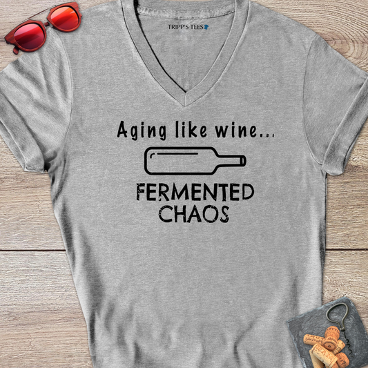 A sport gray v-neck t-shirt with a black distressed graphic reading 'Aging like wine...Fermented Chaos' and a graphic of a wine bottle on it's side, laid flat on a wood surface with sunglasses, wine opener and corks on a coaster. 