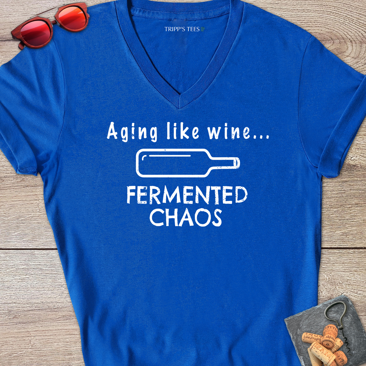 A royal blue v-neck t-shirt with a white distressed graphic reading 'Aging like wine...Fermented Chaos' and a graphic of a wine bottle on it's side, laid flat on a wood surface with sunglasses, wine opener and corks on a coaster. 