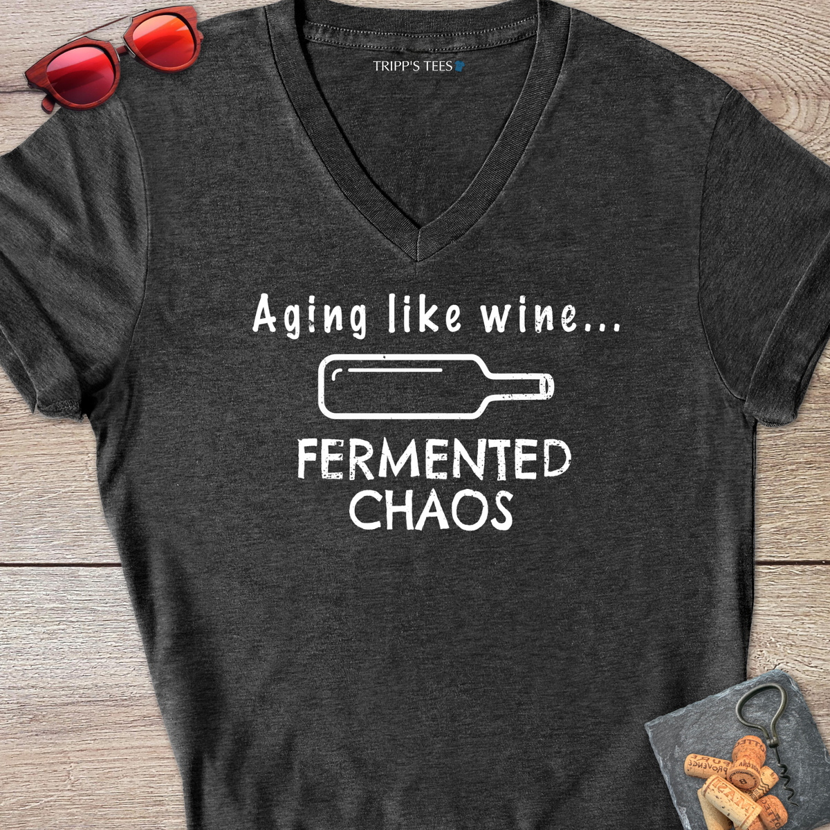 A dark gray heather v-neck t-shirt with a white distressed graphic reading 'Aging like wine...Fermented Chaos' and a graphic of a wine bottle on it's side, laid flat on a wood surface with sunglasses, wine opener and corks on a coaster. 