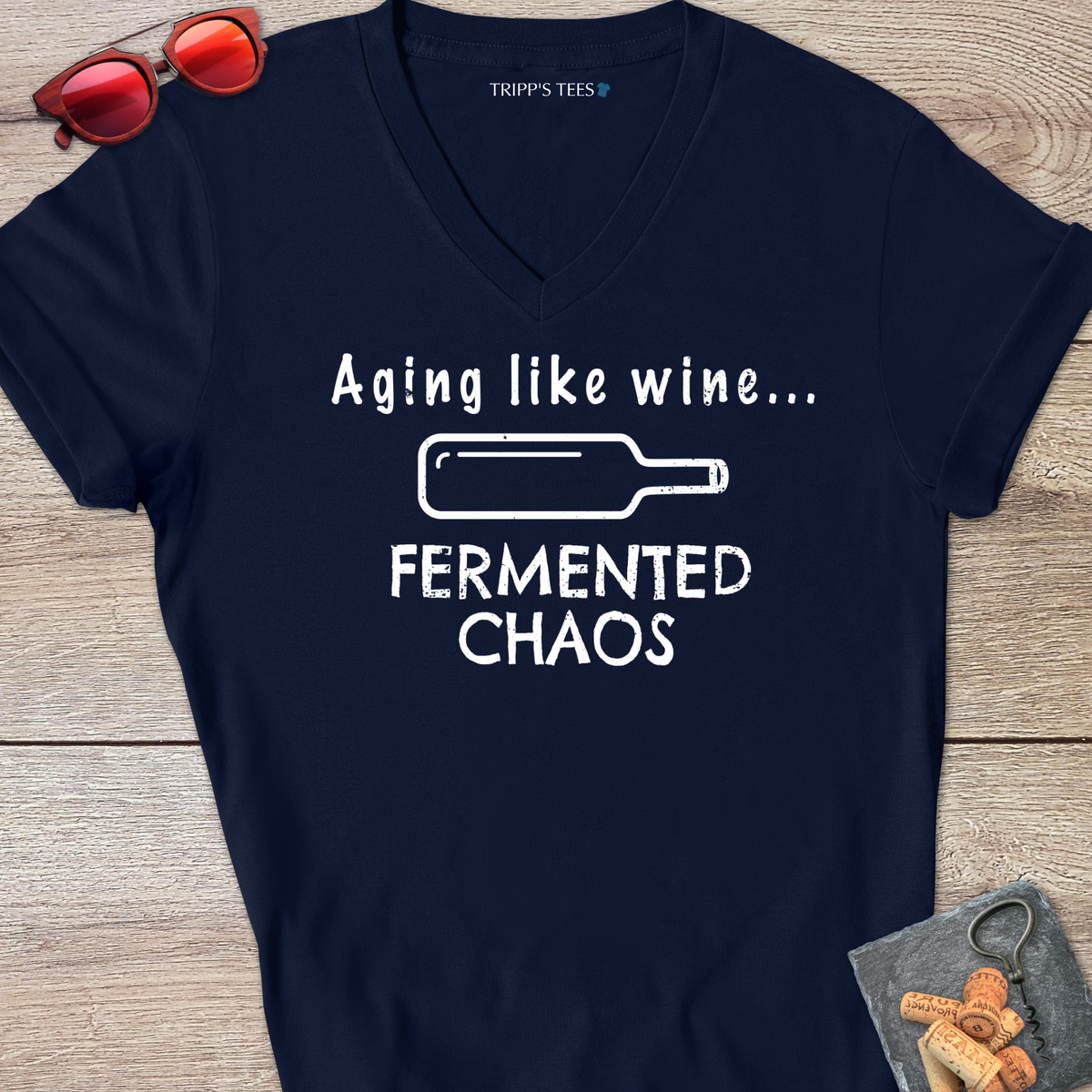 A navy v-neck t-shirt with a white distressed graphic reading 'Aging like wine...Fermented Chaos' and a graphic of a wine bottle on it's side, laid flat on a wood surface with sunglasses, wine opener and corks on a coaster. 