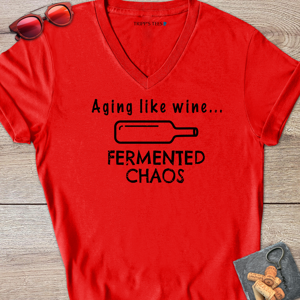 A red v-neck t-shirt with a black distressed graphic reading 'Aging like wine...Fermented Chaos' and a graphic of a wine bottle on it's side, laid flat on a wood surface with sunglasses, wine opener and corks on a coaster. 