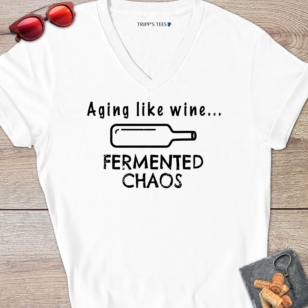 A white v-neck t-shirt with a black distressed graphic reading 'Aging like wine...Fermented Chaos' and a graphic of a wine bottle on it's side, laid flat on a wood surface with sunglasses, wine opener and corks on a coaster. 