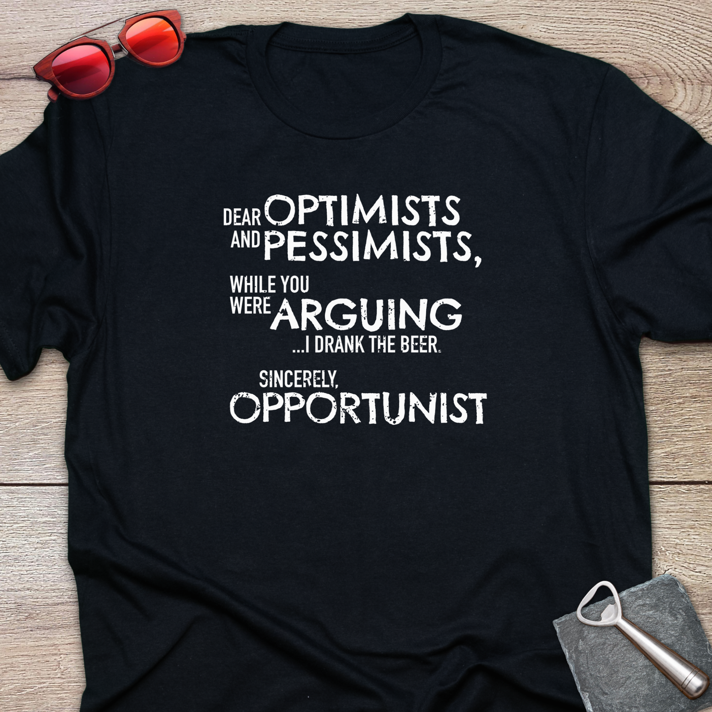 An black graphic t-shirt laid flat on a light wood background, styled with red sunglasses at the top left and a gray bottle opener at the bottom right. The t-shirt features a white printed quote that reads: 'DEAR OPTIMISTS AND PESSIMISTS, WHILE YOU WERE ARGUING...I drank the beer. Sincerely, OPPORTUNIST."