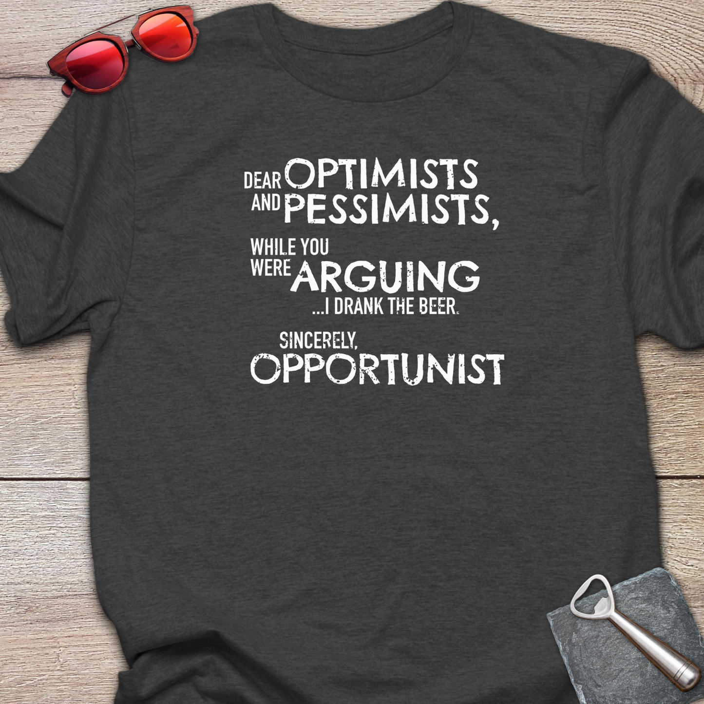 An dark heather graphic t-shirt laid flat on a light wood background, styled with red sunglasses at the top left and a gray bottle opener at the bottom right. The t-shirt features a white printed quote that reads: 'DEAR OPTIMISTS AND PESSIMISTS, WHILE YOU WERE ARGUING...I drank the beer. Sincerely, OPPORTUNIST."