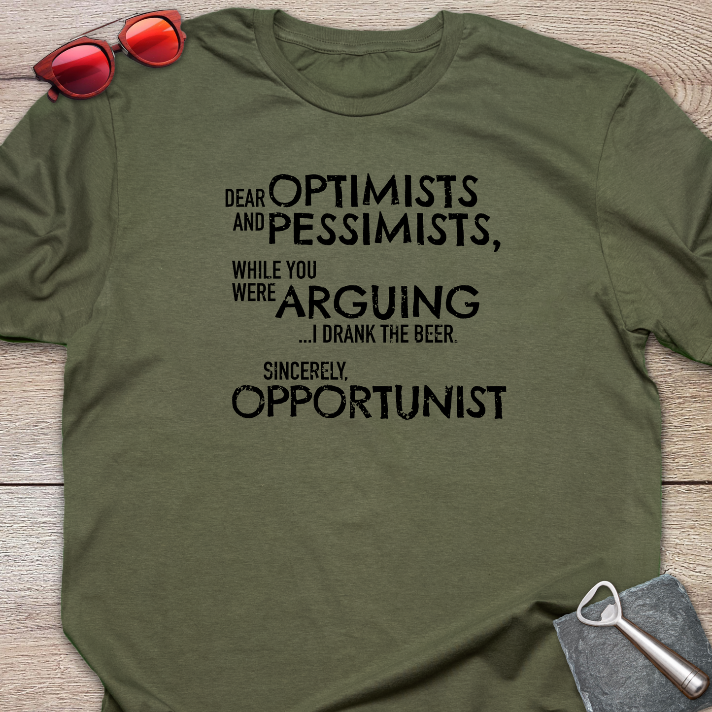 An olive green graphic t-shirt laid flat on a light wood background, styled with red sunglasses at the top left and a gray bottle opener at the bottom right. The t-shirt features a black printed quote that reads: 'DEAR OPTIMISTS AND PESSIMISTS, WHILE YOU WERE ARGUING...I drank the beer. Sincerely, OPPORTUNIST."