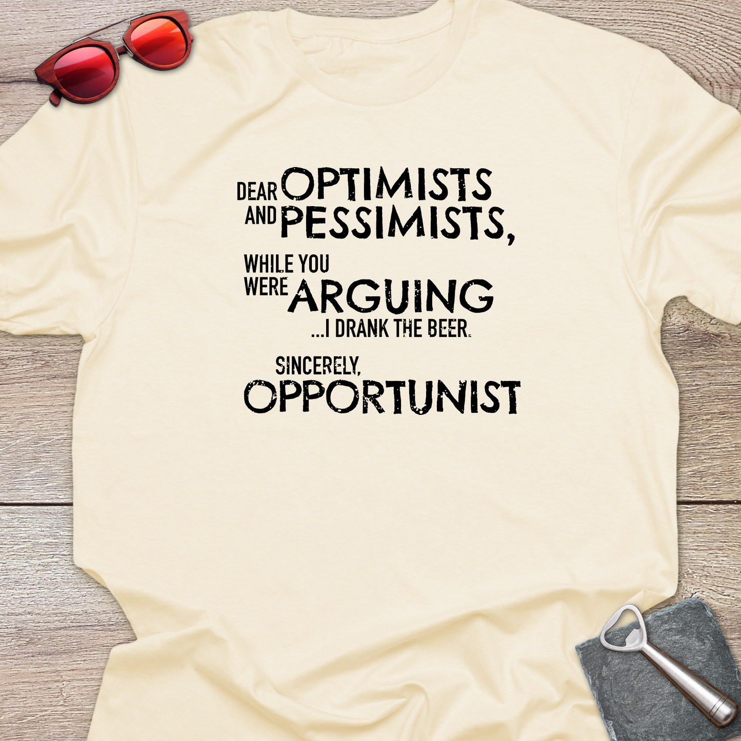 An natural graphic t-shirt laid flat on a light wood background, styled with red sunglasses at the top left and a gray bottle opener at the bottom right. The t-shirt features a black printed quote that reads: 'DEAR OPTIMISTS AND PESSIMISTS, WHILE YOU WERE ARGUING...I drank the beer. Sincerely, OPPORTUNIST."