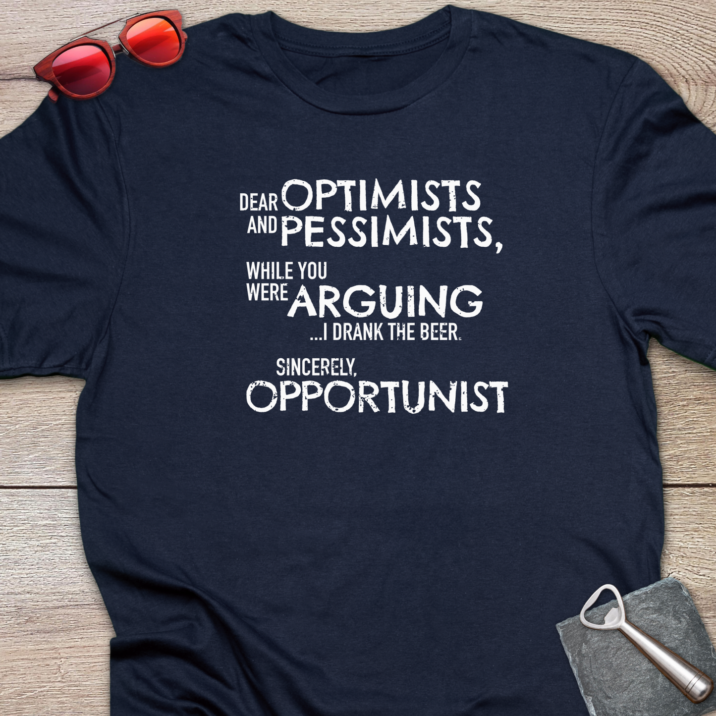 An navy blue graphic t-shirt laid flat on a light wood background, styled with red sunglasses at the top left and a gray bottle opener at the bottom right. The t-shirt features a white printed quote that reads: 'DEAR OPTIMISTS AND PESSIMISTS, WHILE YOU WERE ARGUING...I drank the beer. Sincerely, OPPORTUNIST."