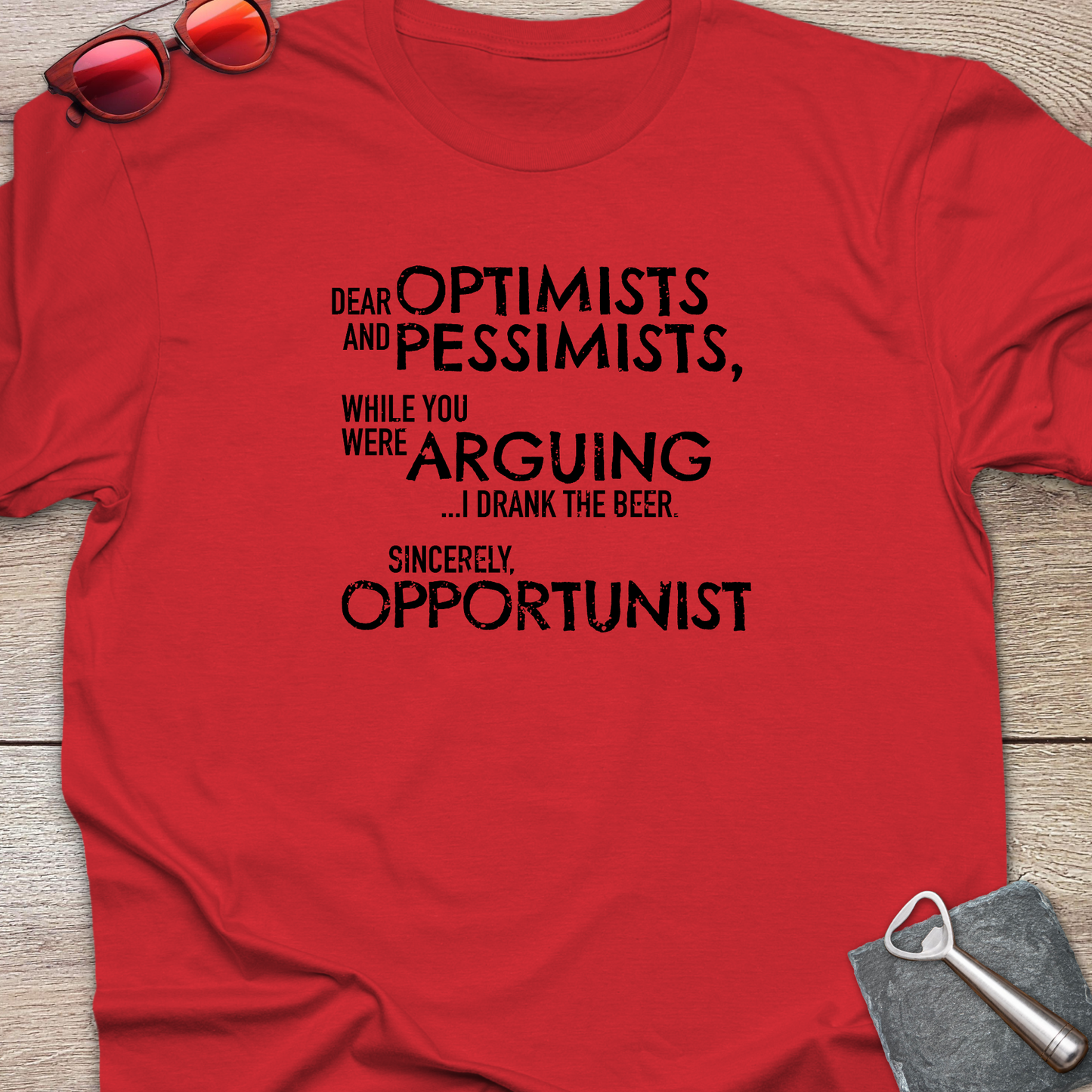 An red graphic t-shirt laid flat on a light wood background, styled with red sunglasses at the top left and a gray bottle opener at the bottom right. The t-shirt features a black printed quote that reads: 'DEAR OPTIMISTS AND PESSIMISTS, WHILE YOU WERE ARGUING...I drank the beer. Sincerely, OPPORTUNIST."