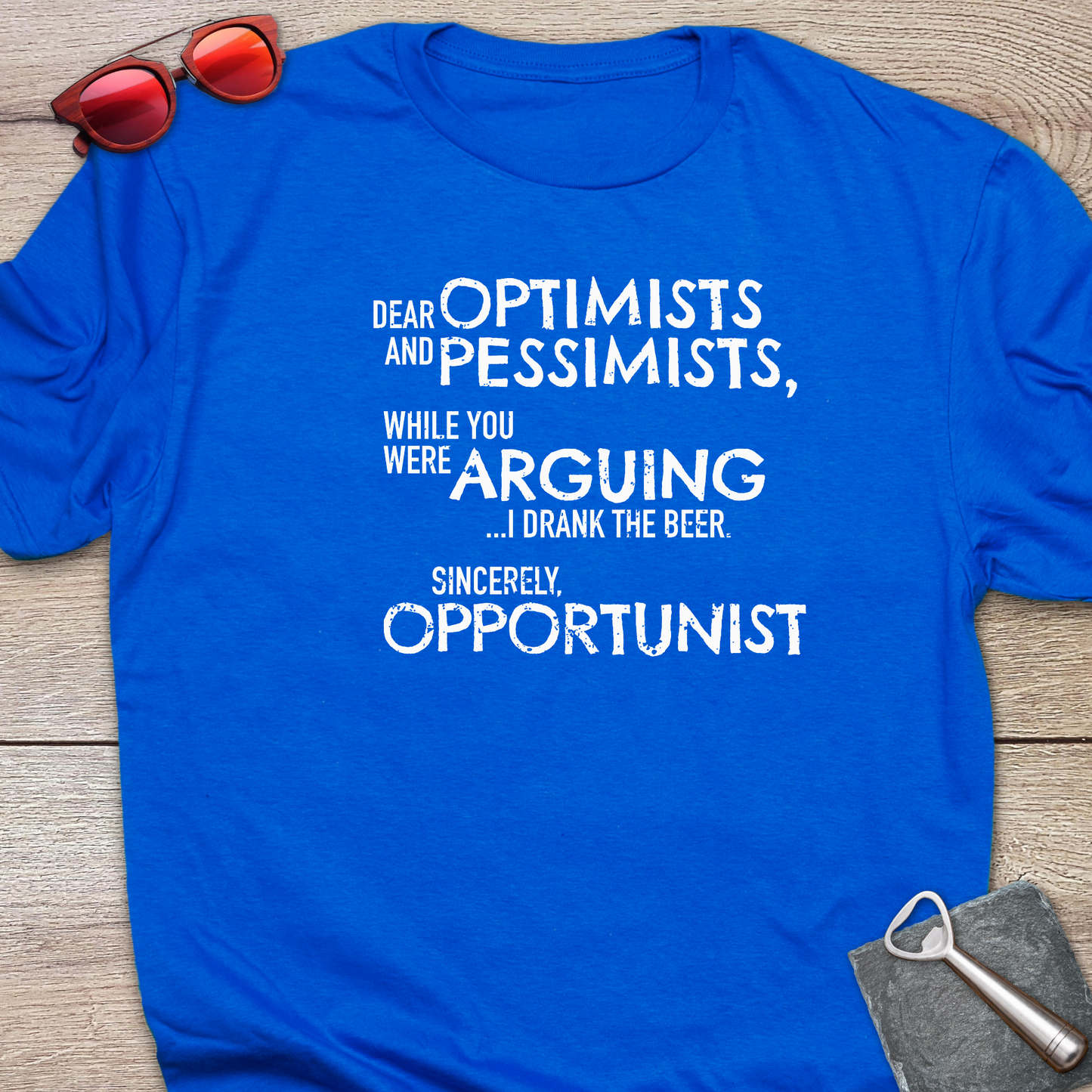 An royal blue graphic t-shirt laid flat on a light wood background, styled with red sunglasses at the top left and a gray bottle opener at the bottom right. The t-shirt features a white printed quote that reads: 'DEAR OPTIMISTS AND PESSIMISTS, WHILE YOU WERE ARGUING...I drank the beer. Sincerely, OPPORTUNIST."