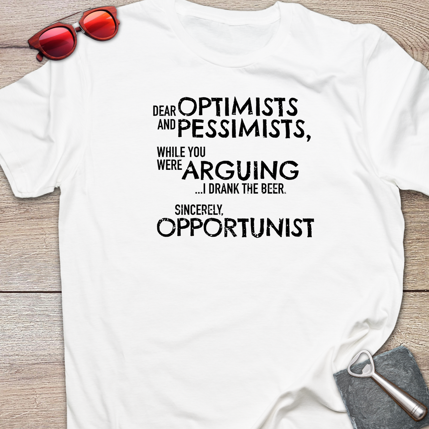 An white graphic t-shirt laid flat on a light wood background, styled with red sunglasses at the top left and a gray bottle opener at the bottom right. The t-shirt features a black printed quote that reads: 'DEAR OPTIMISTS AND PESSIMISTS, WHILE YOU WERE ARGUING...I drank the beer. Sincerely, OPPORTUNIST."