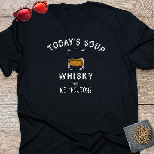 A black short-sleeve graphic T-shirt laid flat on a light wood surface, with red sunglasses resting near the collar and a small coaster with loose hops on it in the lower right corner. The shirt features a white printed slogan reading “TODAY'S SOUP WHISKY WITH ICE CROUTONS” around a small whisky glass graphic.