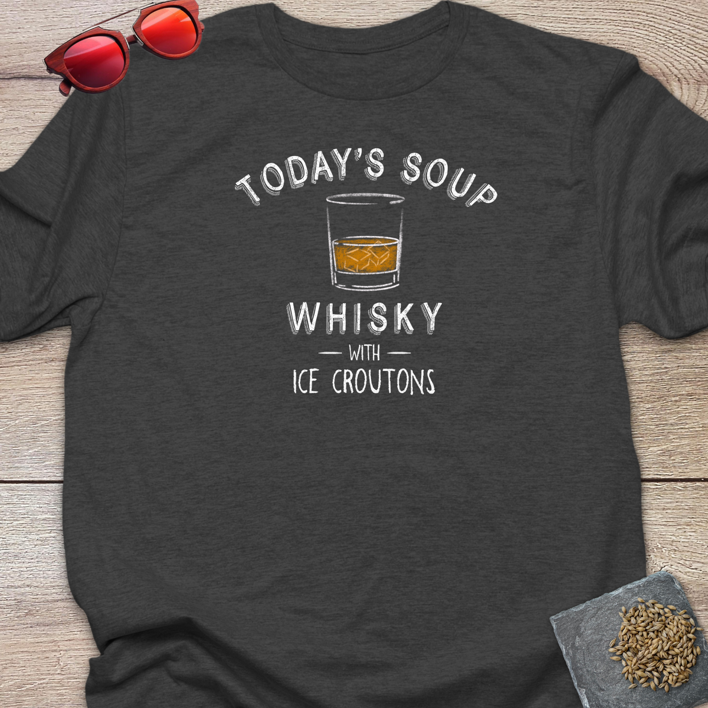 A dark charcoal gray t-shirt laid flat on a light wood background, featuring a white graphic that reads 'TODAY'S SOUP WHISKY WITH ICE CROUTONS' around an illustration of a whiskey glass. Red-tinted sunglasses rest in the top-left corner, and a small coaster with loose hops on it is in the bottom-right corner.