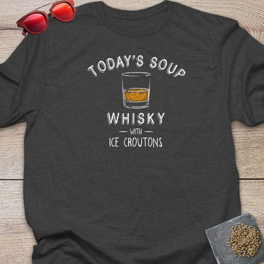 A dark charcoal gray t-shirt laid flat on a light wood background, featuring a white graphic that reads 'TODAY'S SOUP WHISKY WITH ICE CROUTONS' around an illustration of a whiskey glass. Red-tinted sunglasses rest in the top-left corner, and a small coaster with loose hops on it is in the bottom-right corner.