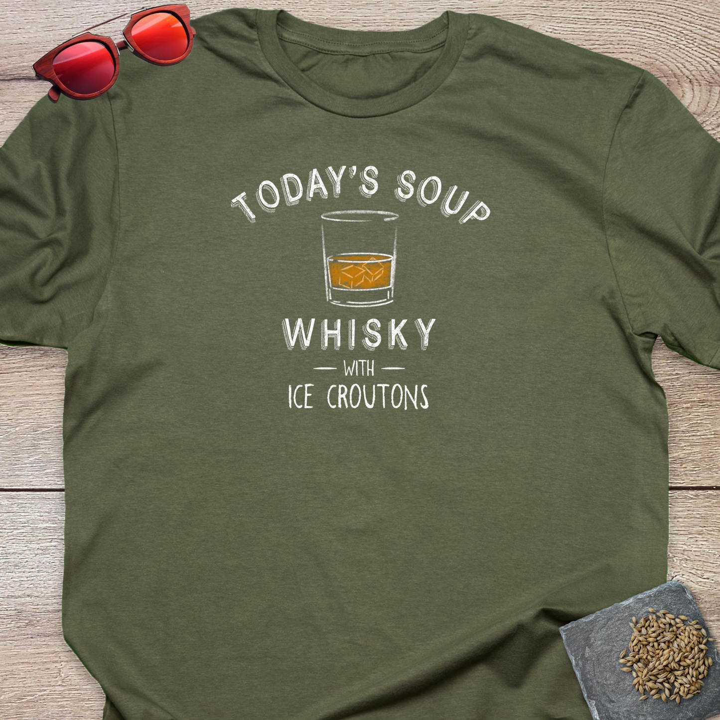 An olive green crewneck t-shirt with a white graphic print that reads 'TODAY'S SOUP WHISKY WITH ICE CROUTONS' and features an illustration of a whiskey glass. The shirt is styled in a flat-lay on a wooden surface with red sunglasses at the top left and a small coaster with loose hops on it  at the bottom right.