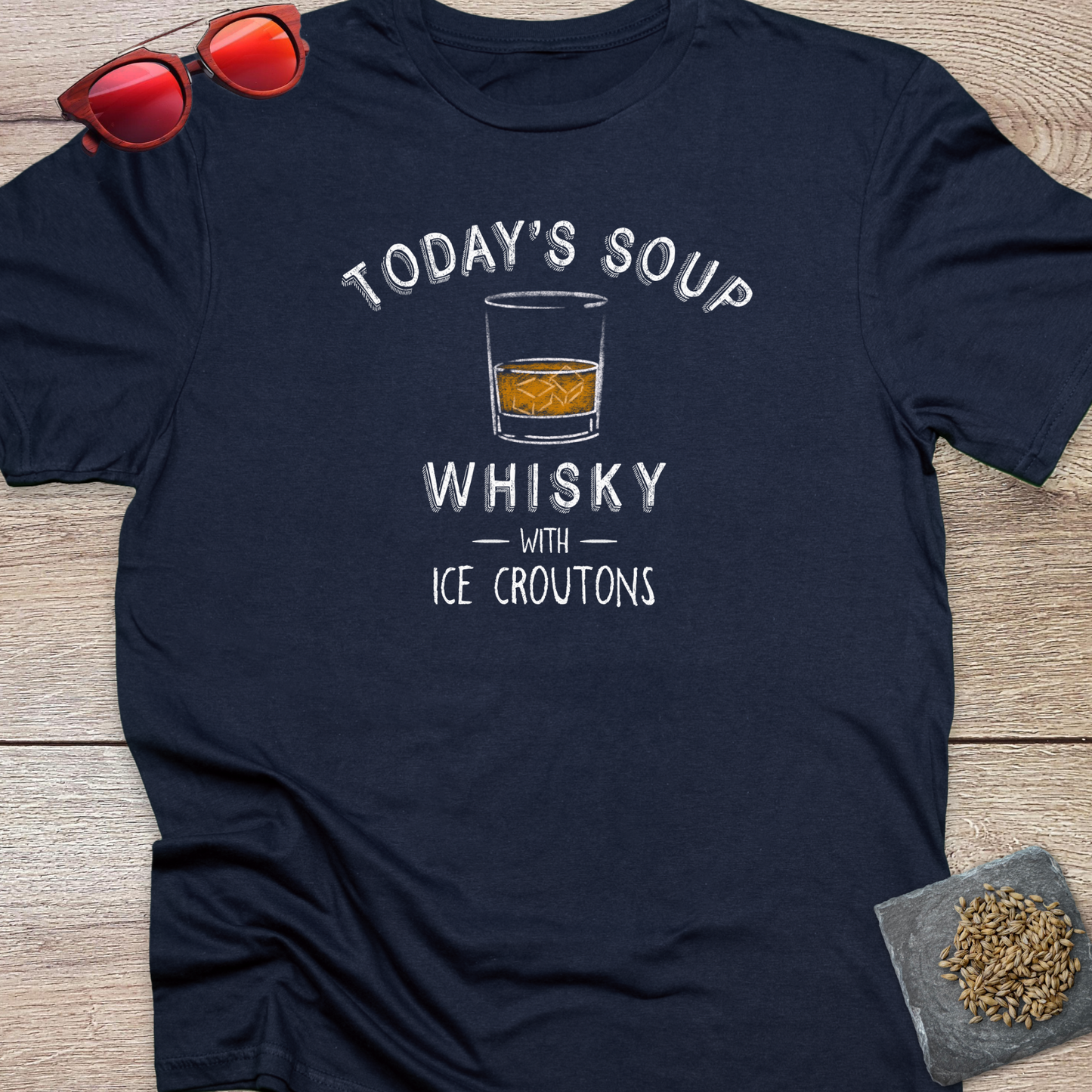 A navy blue graphic t-shirt laid flat on a wooden surface, featuring a white printed design that reads 'TODAY'S SOUP WHISKY WITH ICE CROUTONS' around an illustration of a whiskey glass. Red-tinted sunglasses are placed in the top-left corner, and a small coaster with some hops on it is in the bottom-right corner.
