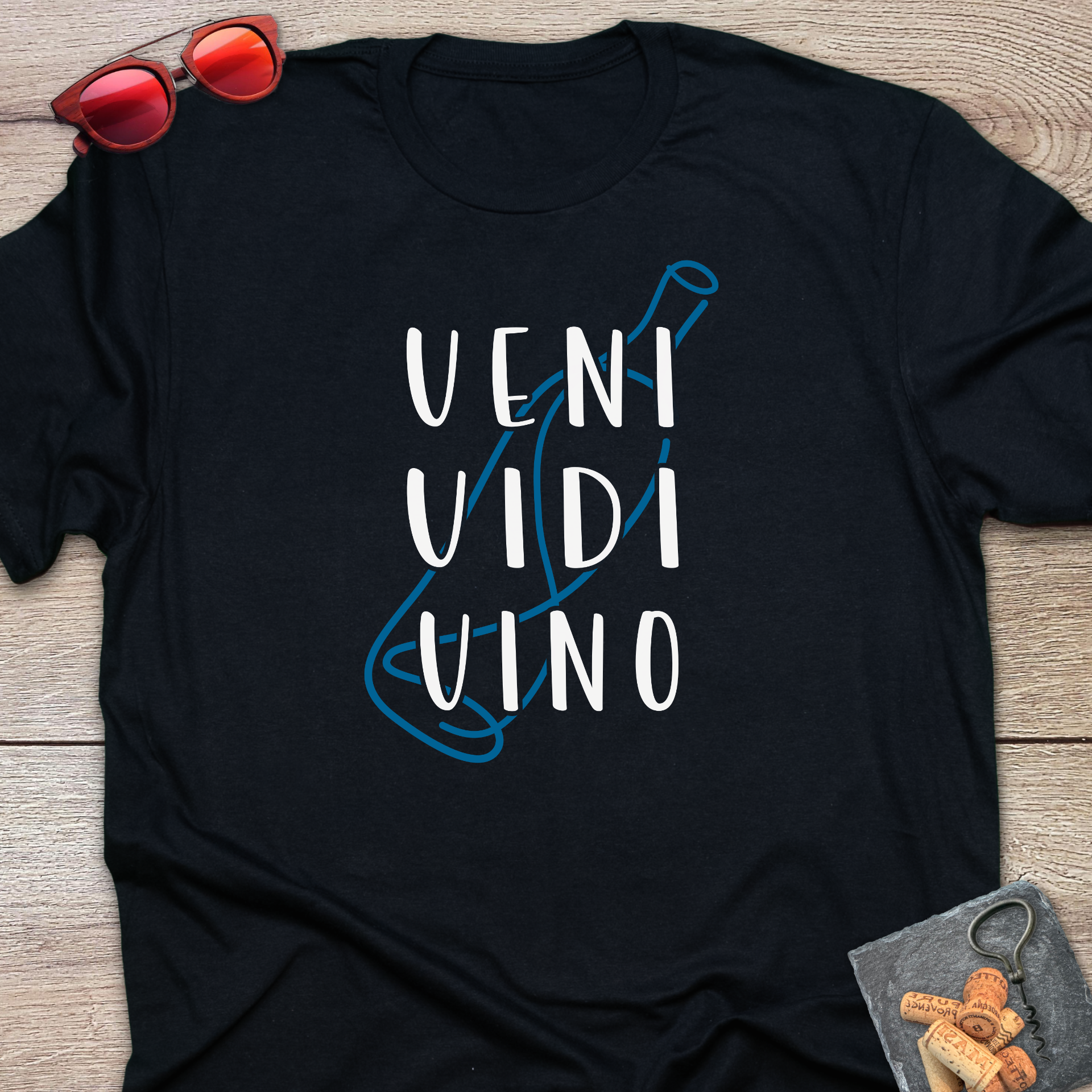 A black short-sleeve t-shirt laid flat on a light wood surface, featuring the phrase 'VENI VIDI VINO' in white block letters over a blue wine bottle outline. Red sunglasses rest at the top left, and a small corkscrew with corks is at the bottom right.