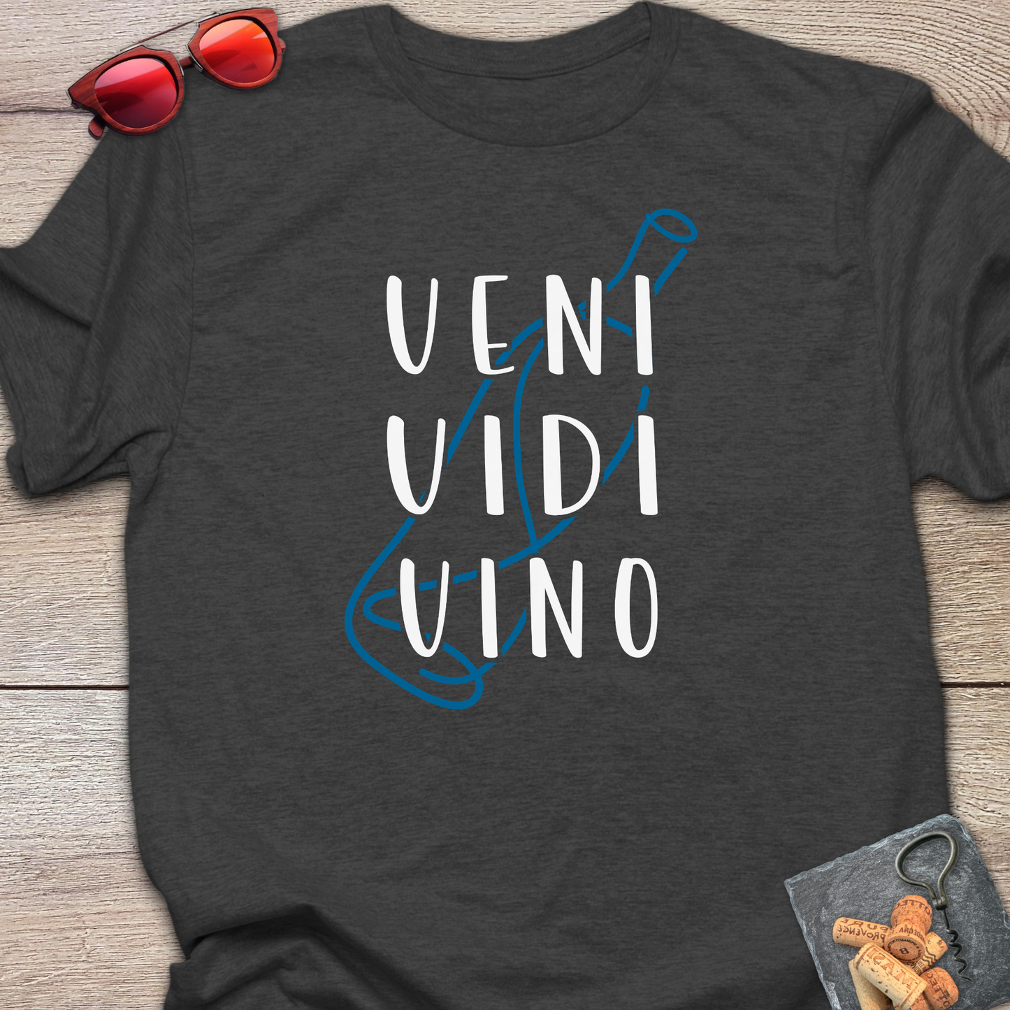A dark charcoal gray t-shirt laid flat on a light wood background, featuring a graphic of a blue wine bottle outline with the text 'VENI VIDI VINO' in white. Styled flat-lay props include red sunglasses in the top-left corner and a corkscrew with corks in the bottom-right corner.