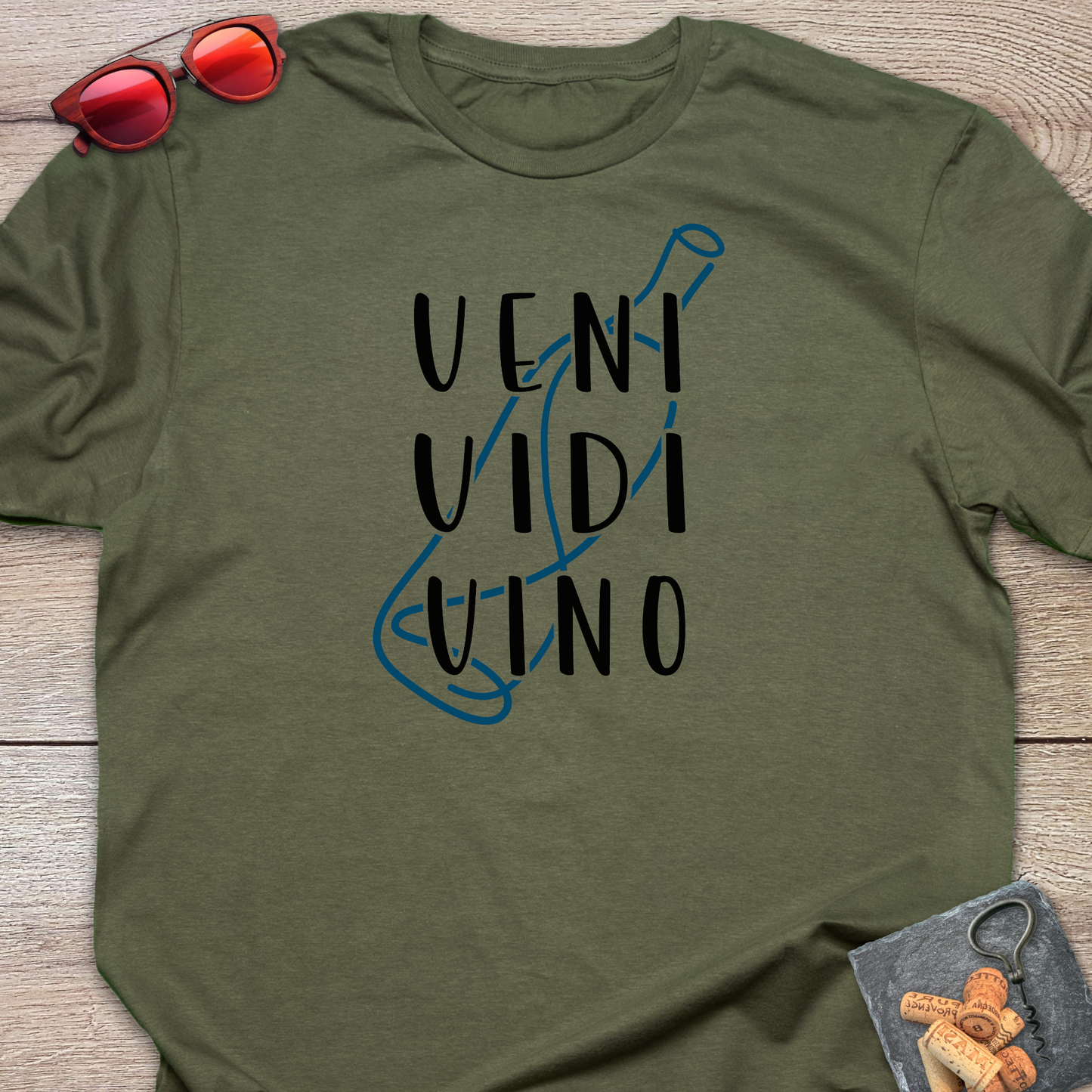 An olive green crewneck t-shirt with a graphic print reading 'VENI VIDI VINO' in black over a blue wine bottle outline, styled flat on a wooden surface with red sunglasses and a small corkscrew and corks in the corners.