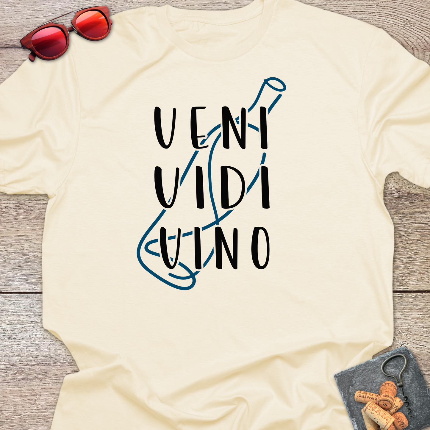 A cream-colored t-shirt laid flat on a gray wooden surface, featuring a graphic print of the words 'VENI VIDI VINO' in black next to a blue wine bottle outline. Styled flat-lay props include red sunglasses at the top left and a small corkscrew with wine corks at the bottom right.