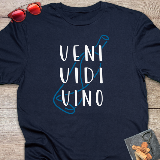 A navy blue t-shirt laid flat on a light wood background, featuring a graphic of a wine bottle in blue outline with the words 'VENI VIDI VINO' printed in white. Red sunglasses and a small corkscrew are styled as props near the shirt.