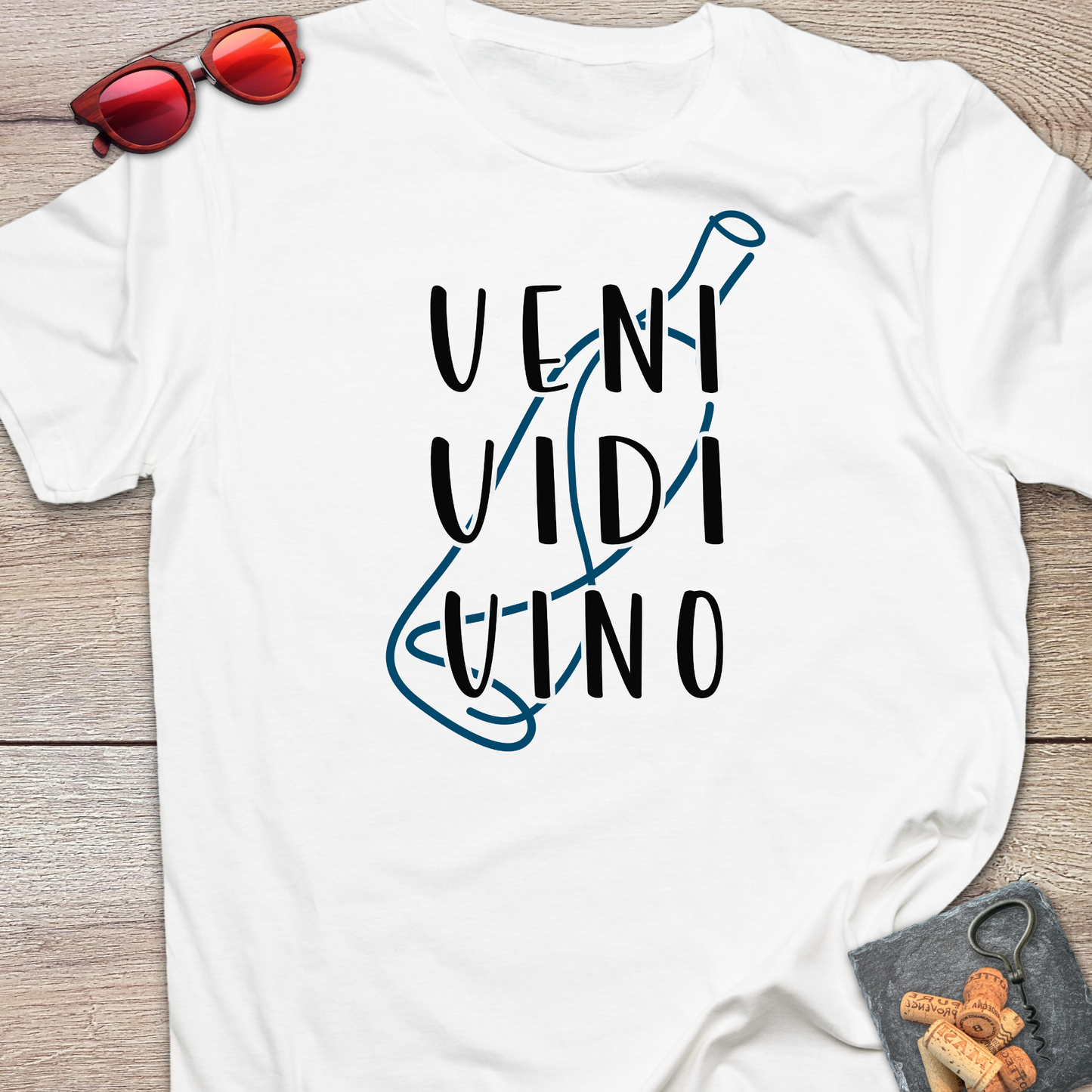 A white short-sleeve t-shirt laid flat on a light wood surface, featuring the text 'VENI VIDI VINO' in black with a blue wine glass outline. Red sunglasses rest in the top-left corner and a small corkscrew and corks in the bottom-right.